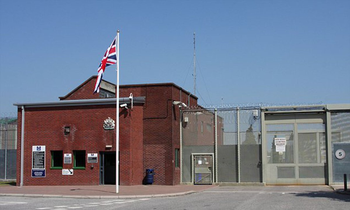 Exterior of a UK prison — red brick gatehouse with Union Jack and perimeter fencing